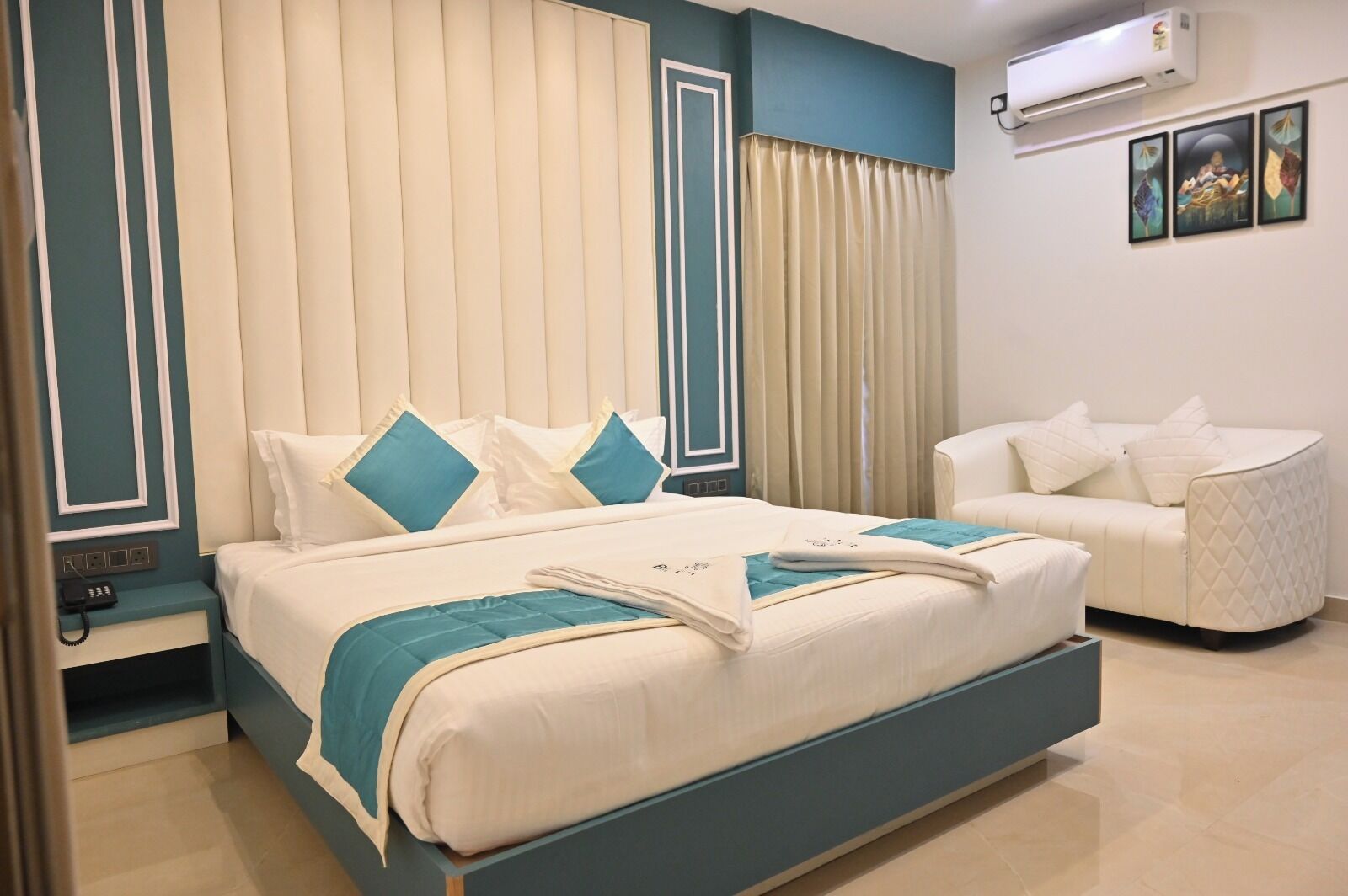A hotel room with a bed, a sofa, a bedside table with a phone, and an air conditioning unit on the wall.