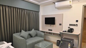 Una habitación de hotel moderna con un sofá gris, un televisor de pantalla plana, una estación de café y un refrigerador pequeño.