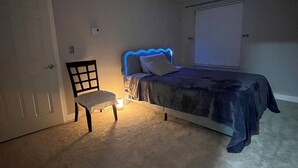Un dormitorio con cama, una silla, mesita de noche, ventana con persianas y una puerta.