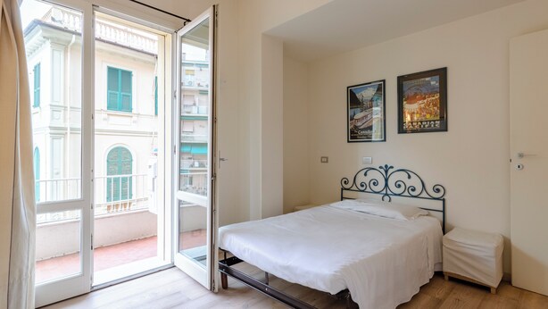 Un dormitorio con cama, mesita de noche pequeña, una puerta y balcón con vistas a edificios.