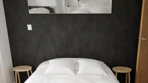 Ein ordentlich gemachtes Bett mit grauer Decke und Kissen, zwei hölzerne Hocker und ein großes gerahmtes Bild an der Wand.