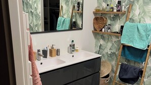 Ein Badezimmer mit weißem Waschbecken, Spiegel und Waschtisch mit dunklem Schrank. Im Boden befindet sich eine Holzleiter mit Handtüchern, einem gewebten Korb und einem grauen Teppich.