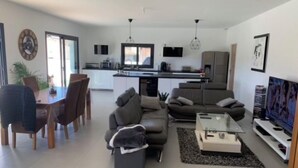 Ein modernes Wohnzimmer mit Sofa, Couchtisch und Fernseher. Ein Essbereich mit Stühlen und Tisch ist ebenfalls vorhanden. Im Hintergrund ist die Küche erkennbar.