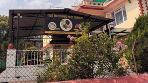 A covered outdoor area with a signboard advertising local cuisine, a fenced garden with flowering plants, and a building with a red roof and beige walls. - 555 Home Stay and Restaurant (Burhanilkantha)