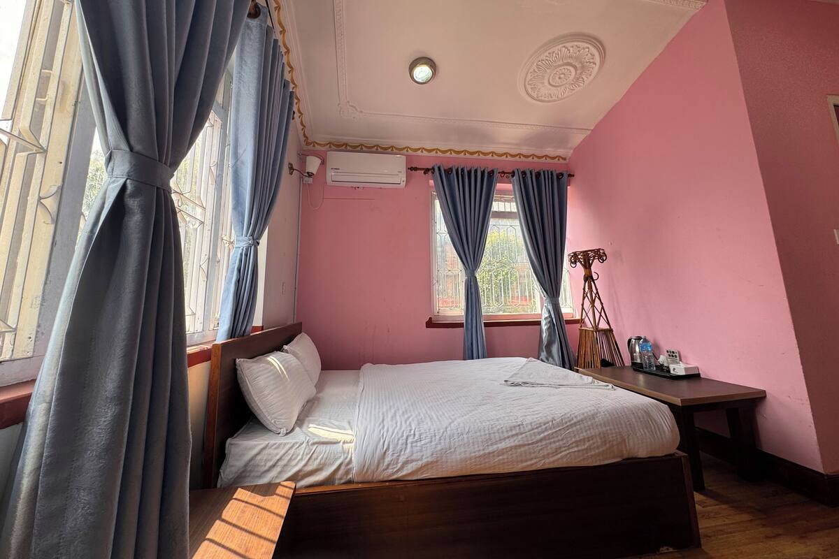 A bedroom with a bed, curtains, an air conditioning unit, a bedside table, and a window.