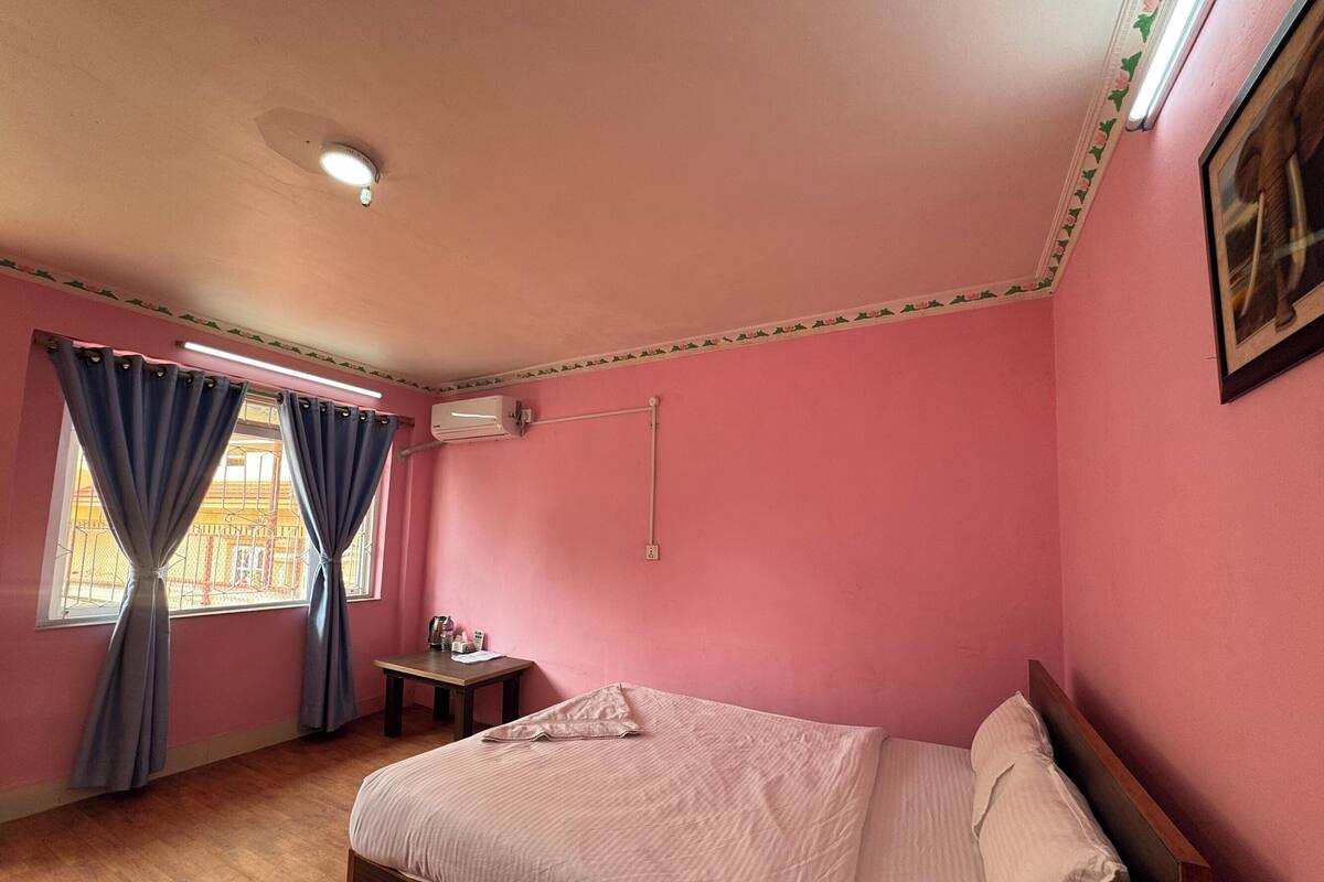 A bedroom with pink walls, a wooden bed, a small bedside table, a window with curtains, and an air conditioning unit.