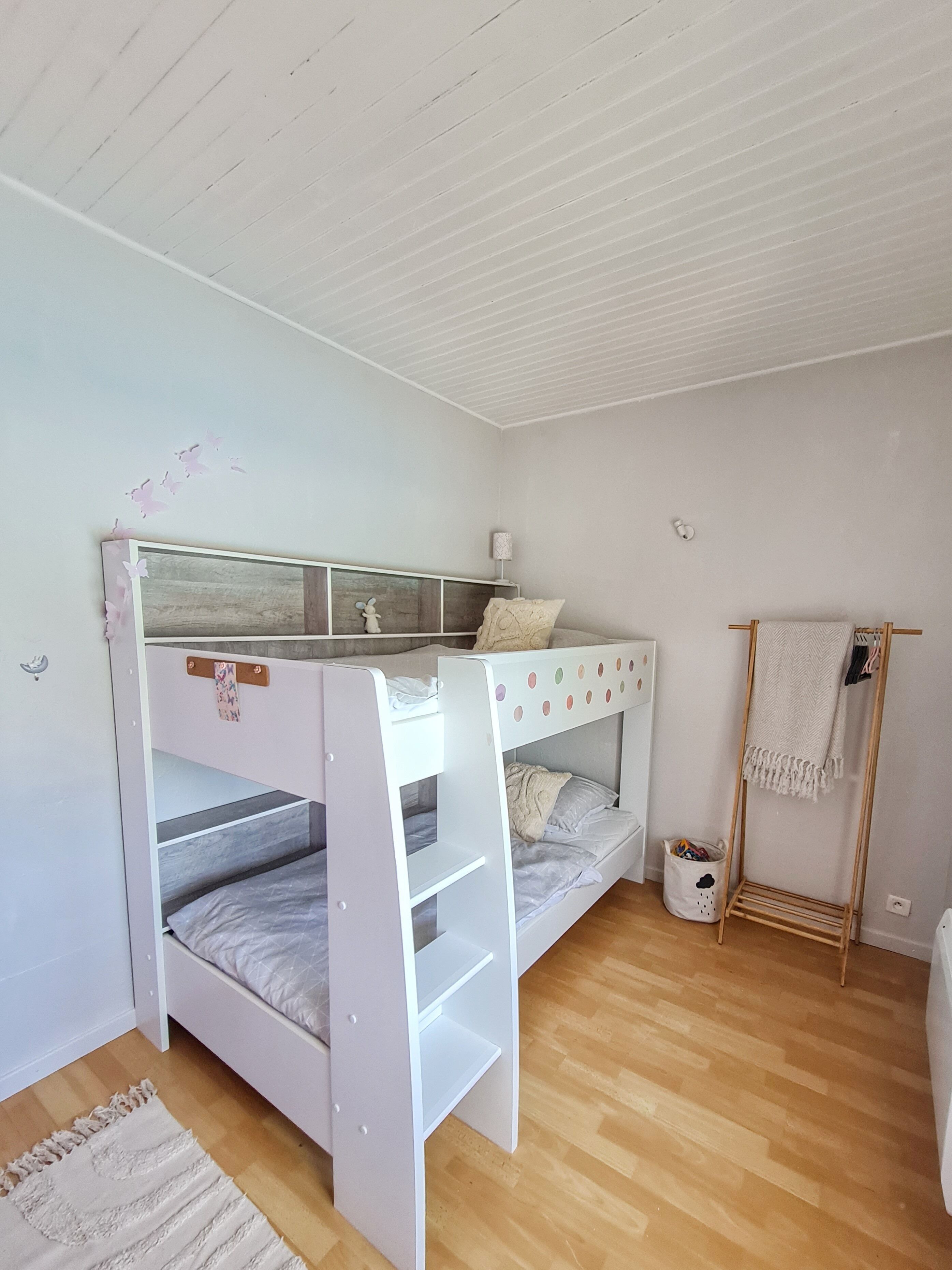 Un lit superposé avec une échelle en bois, une petite table de chevet avec une lampe, un mur blanc et un plancher en bois.