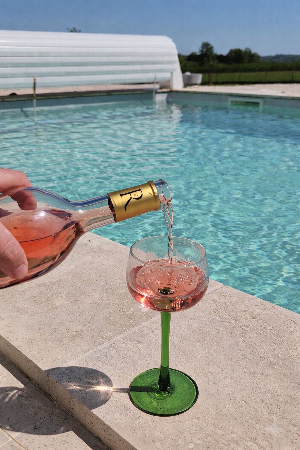 A glass of pink liquid being poured from a bottle with a gold cap into a green glass with a stem.