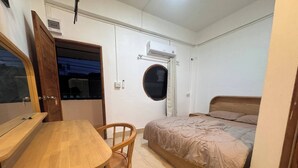 Ein kleines Zimmer mit Holztisch, einem Einzelbett, einem Stuhl, einem Rundfenster und einer Tür.