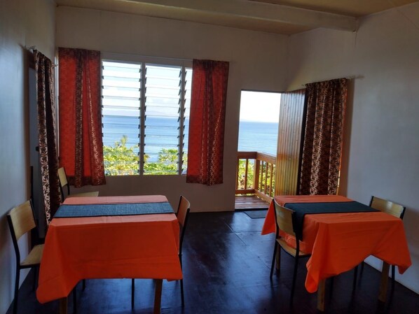 A room with two tables, each with a tablecloth, and a view of the ocean through large windows.