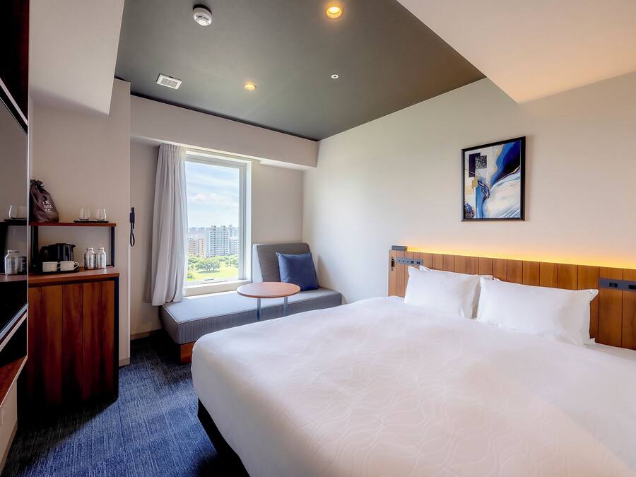 A hotel room with a large bed, a seating area with a view, a small table, and a cabinet with items.