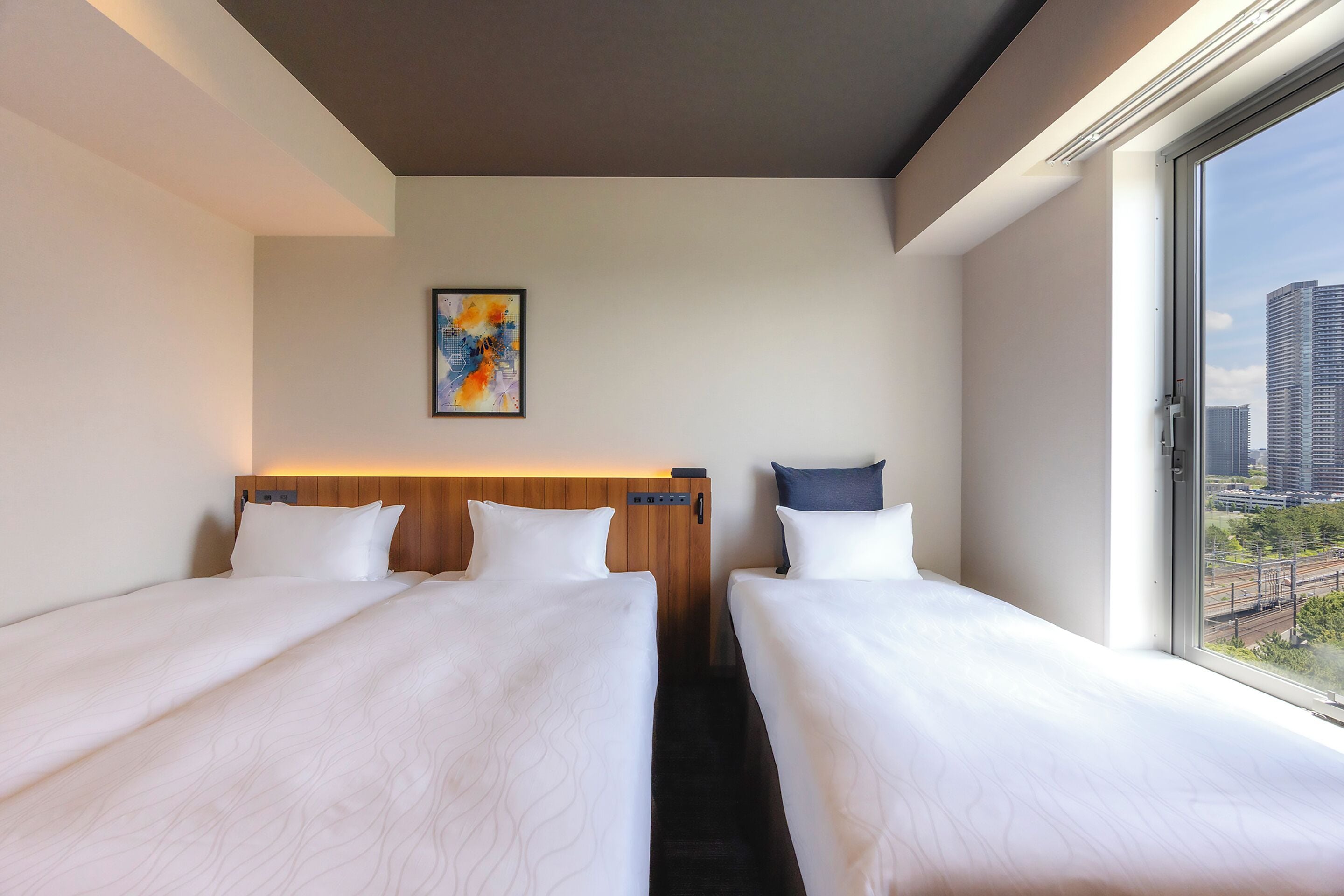 A hotel room with two beds, a wooden headboard, a painting, and a view of a cityscape through a large window.