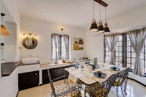 Un comedor con una mesa preparada para cuatro personas, un ventanal con cortinas, un espejo redondo y una encimera de cocina con fregadero.