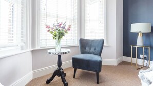 Ein Zimmer mit blauem Sessel, einem kleinen Tisch mit Blumenvase und einer Lampe auf einem Beistelltisch.