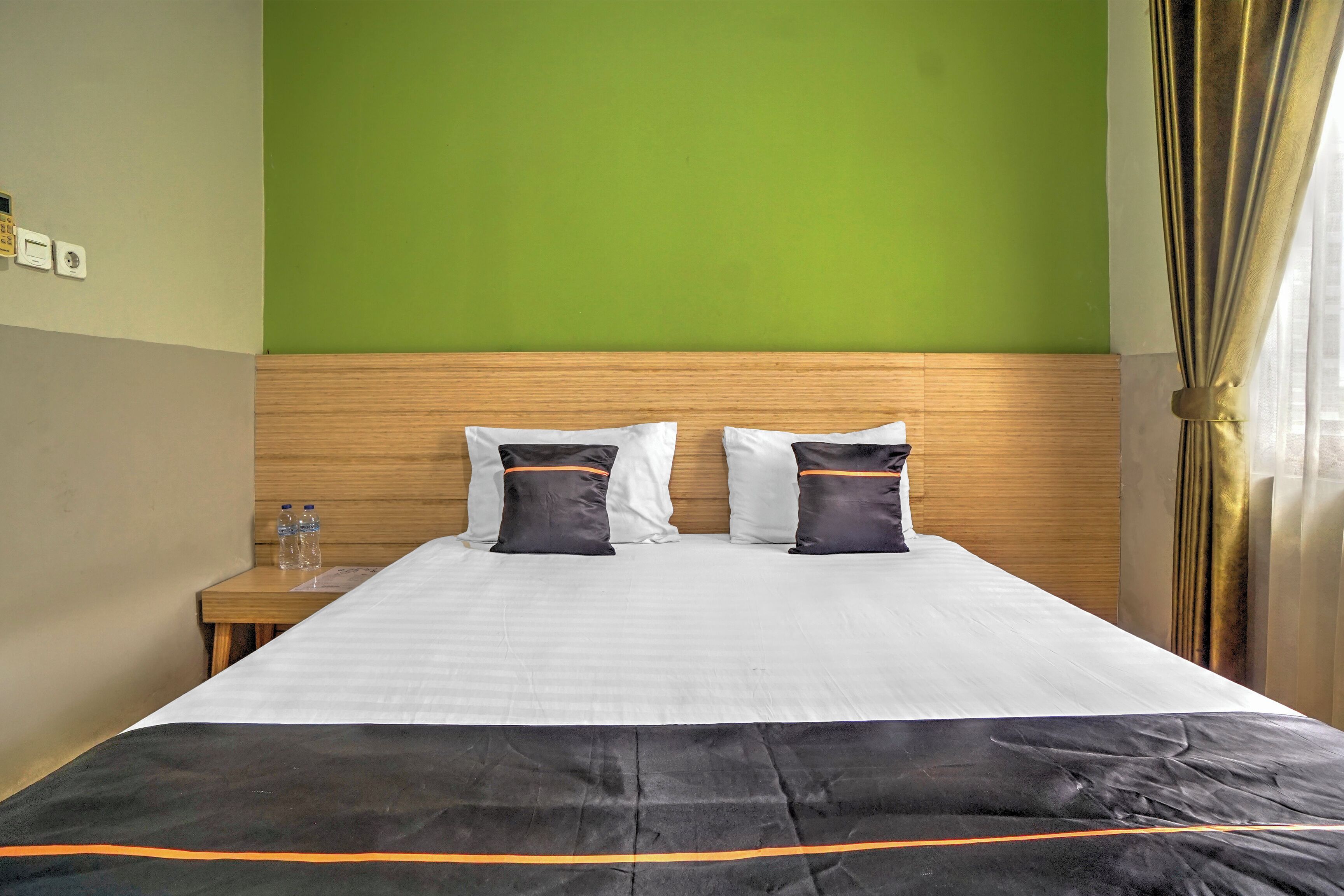 A hotel room with a bed, two pillows, a wooden headboard, a nightstand with a bottle, and a green wall.