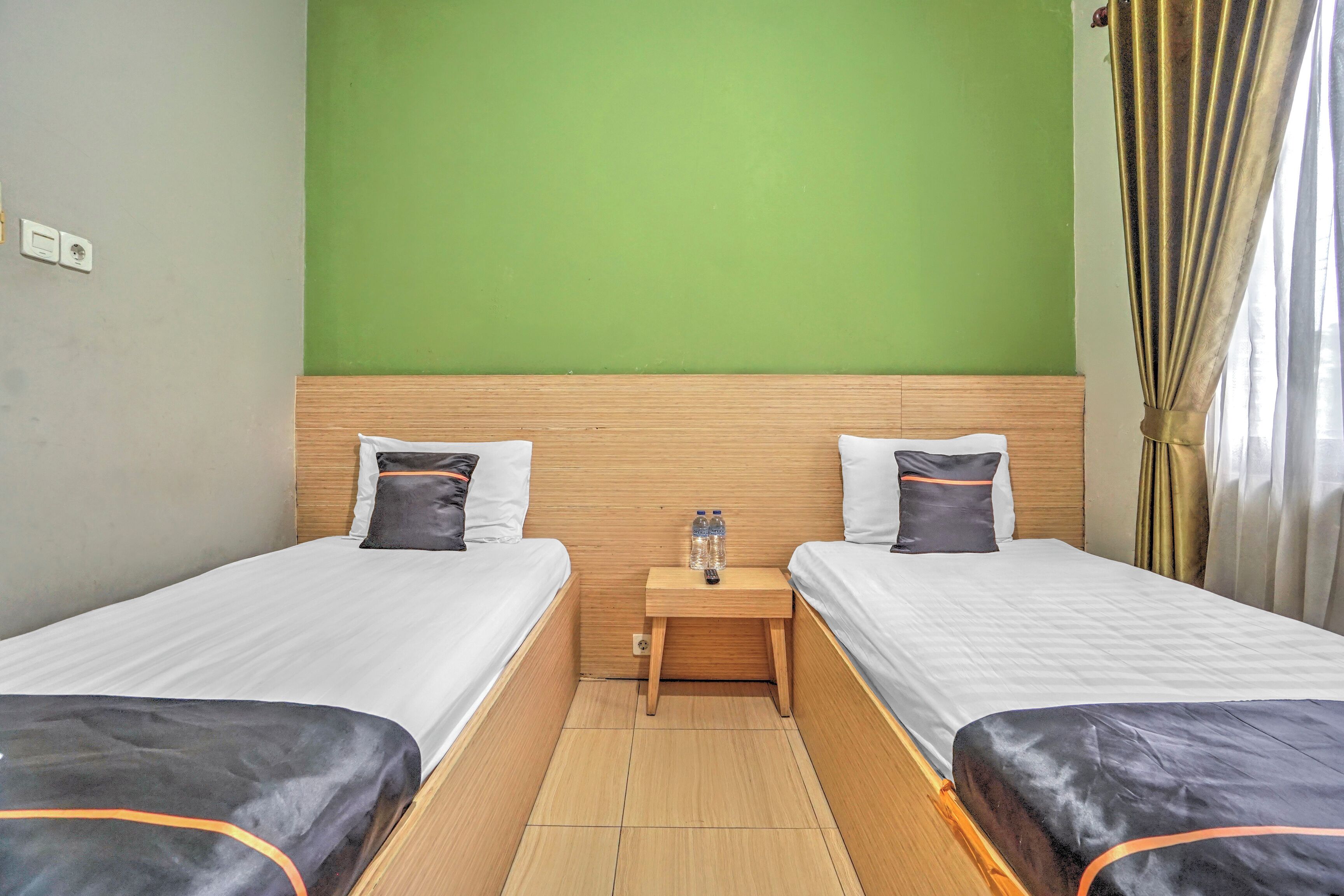 A hotel room with two beds, a wooden headboard, a small bedside table with water bottles, and a green wall.