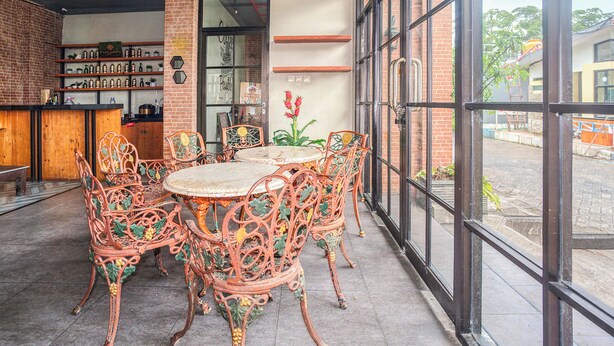 A dining area with a round table and ornate chairs, a brick wall, shelves with bottles, and a view through large windows. - Hotel O near Juanda Airport formerly Azz (Sidoarjo)