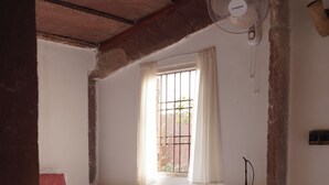 A room with a bed, a window with curtains, a standing fan, and a wall-mounted lamp.
