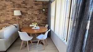 A dining area with a brick wall, a round wooden table, white chairs, and a window with curtains.