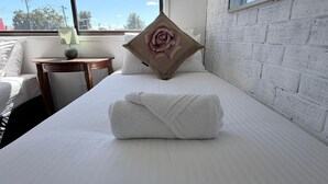 A hotel room with a neatly made bed, a pillow with a rose design, a bedside table with a lamp, and a view of the outdoors through a window.