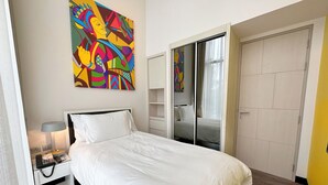 Une chambre d’hôtel comprenant un lit, un téléphone sur la table de chevet, une lampe, un tableau abstrait aux couleurs vives, une armoire et une porte coulissante donnant accès à la salle de bain.