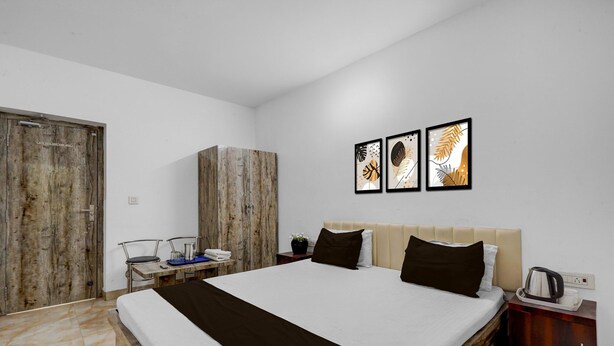 A bedroom with a bed, a wooden wardrobe, a nightstand, a coffee maker, and three framed artworks on the wall.