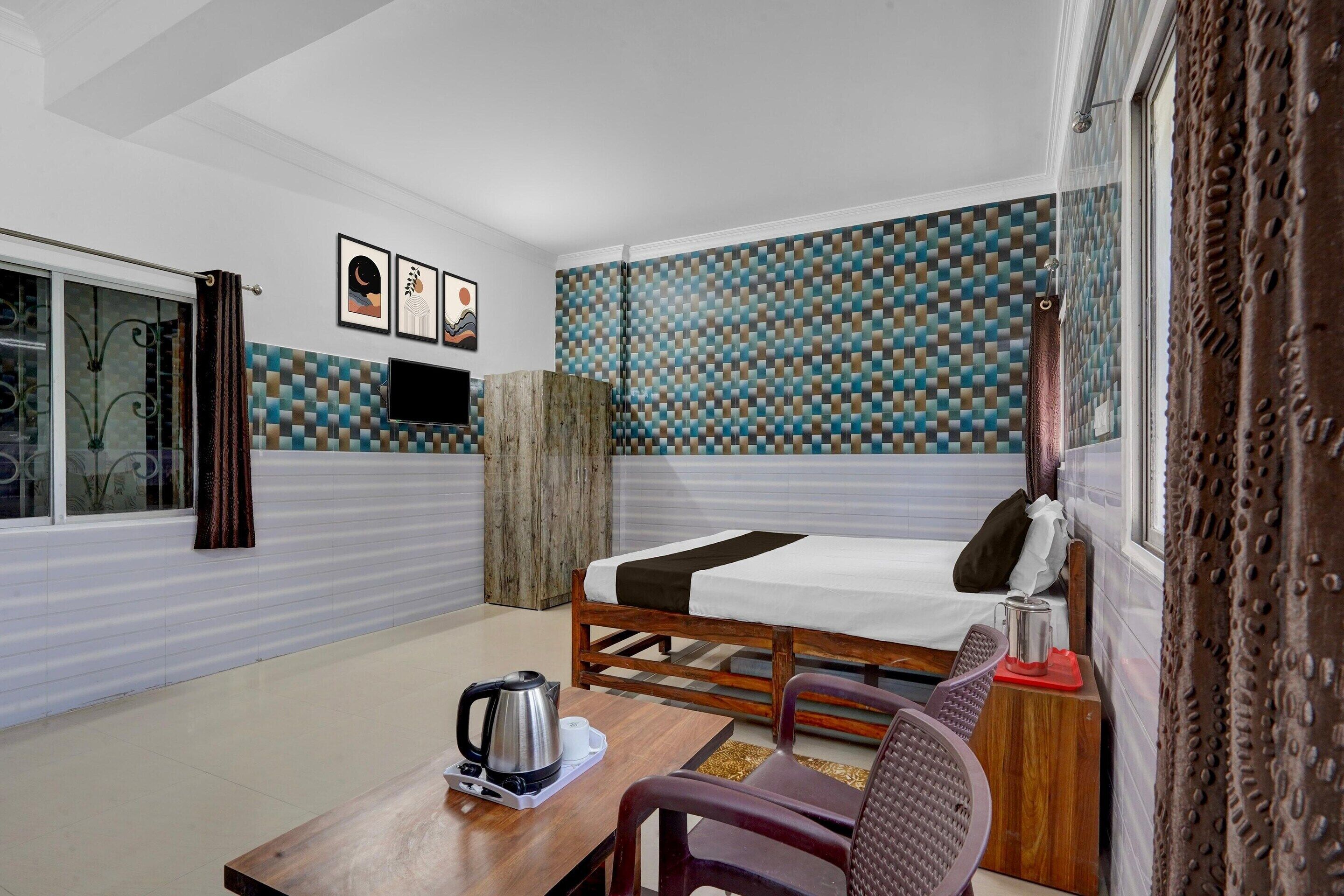 A room with a bed, a table with a kettle, a chair, a TV, and a patterned wall.