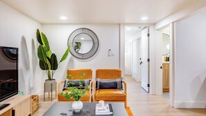 Ein modernes Wohnzimmer mit Fernseher, zwei Sesseln, einem Couchtisch, einer großen Pflanze und einem wandmontierten Spiegel.