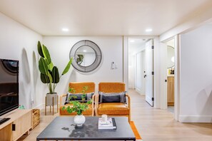 A modern living room with a TV, two armchairs, a coffee table, a large plant, and a wall-mounted mirror. - AC / New Remodel / In-Unit Laundry / Cap Hill (Seattle)
