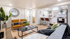 Ein modernes Wohnzimmer mit Sofa, Sessel, Couchtisch und einem Essbereich mit Stühlen sowie einer Küche im Hintergrund.