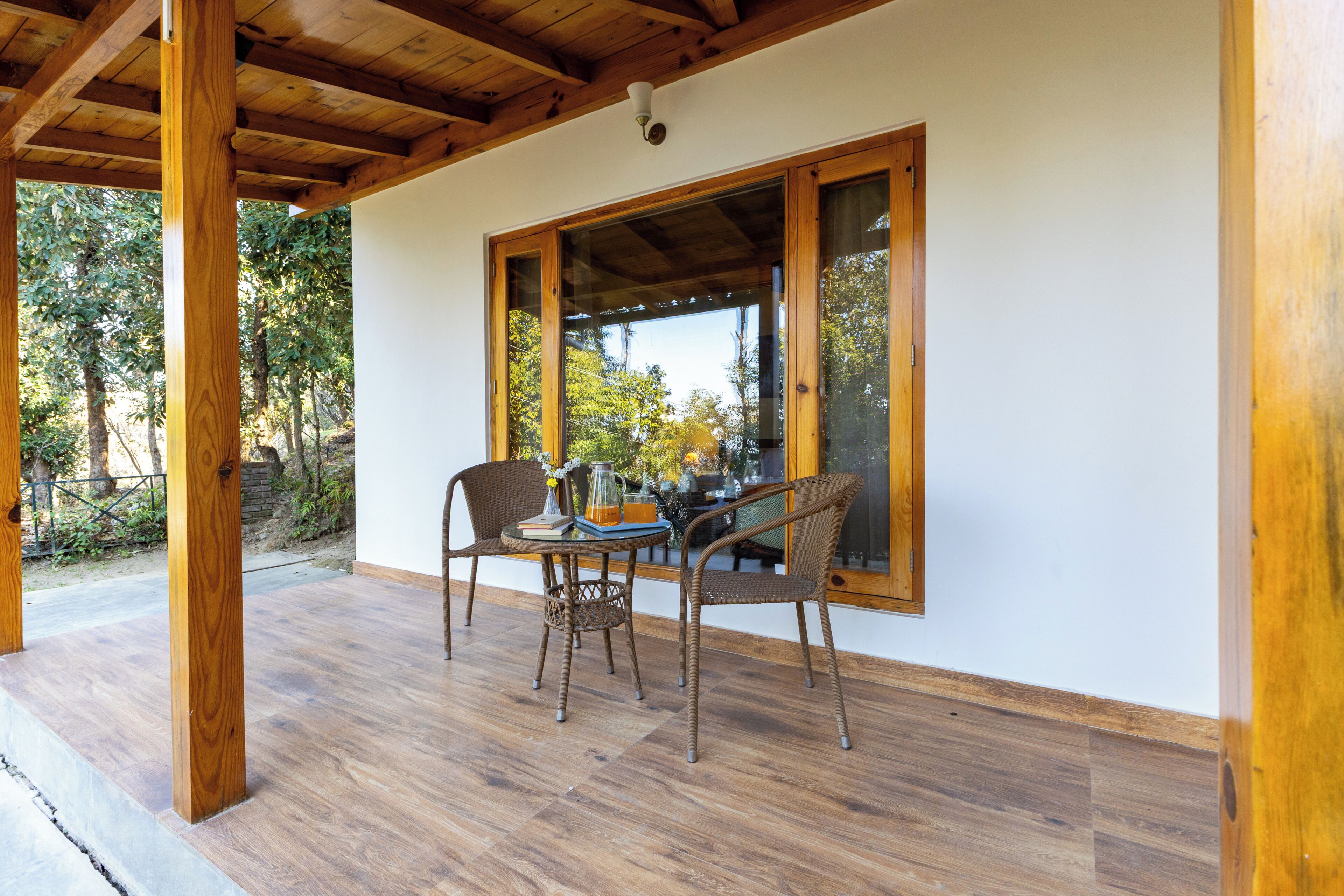 Un patio al aire libre con piso de madera, dos sillas de mimbre, una mesa con un florero y libros, y un ventanal con vista a árboles.