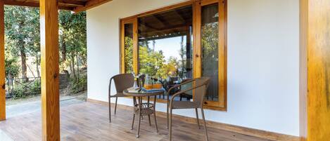 Un patio al aire libre con piso de madera, dos sillas de mimbre, una mesa con un florero y libros, y un ventanal con vista a árboles.