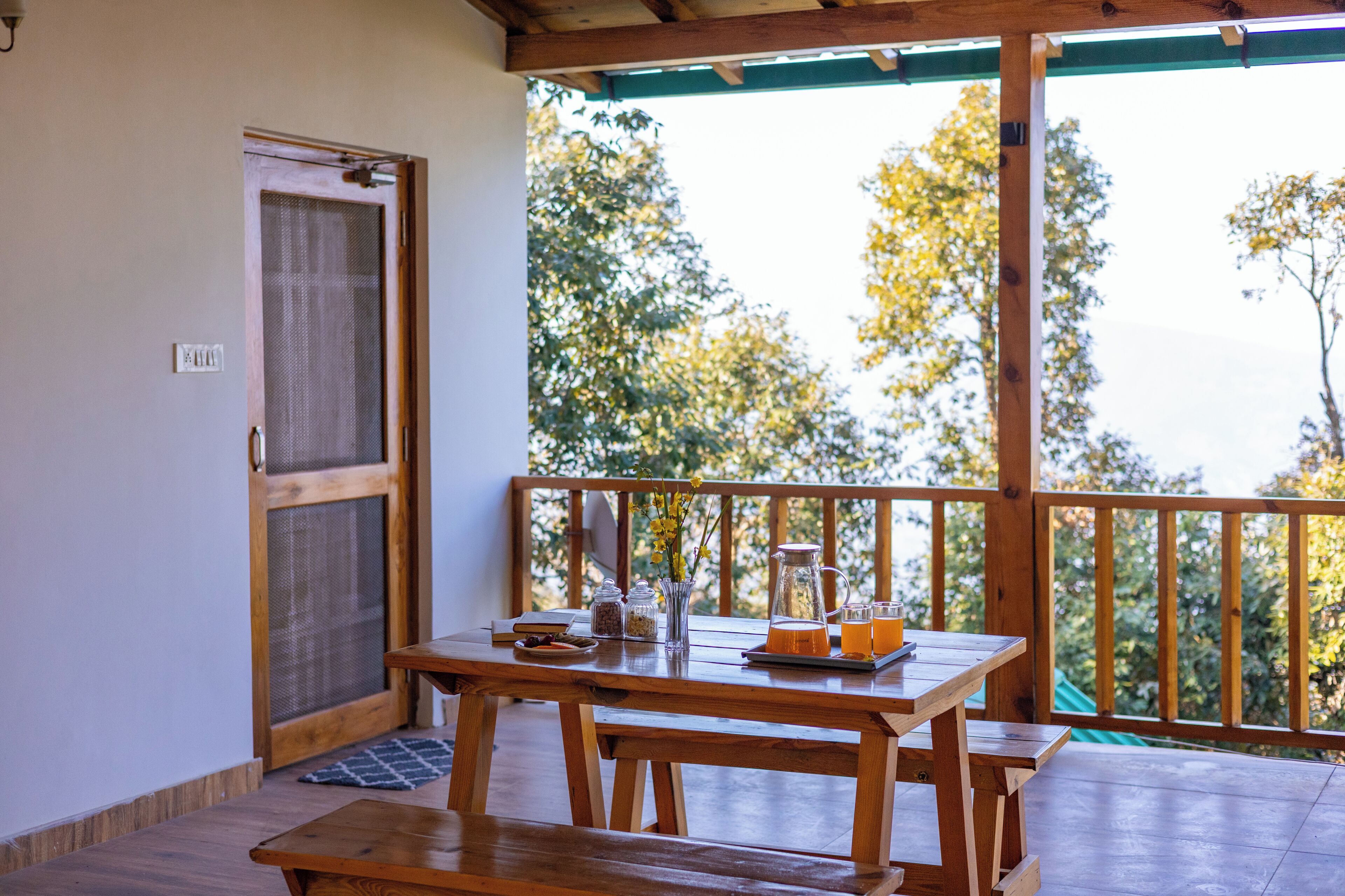 A wooden table with benches, a door with a screen, and a view of trees through a balcony railing.