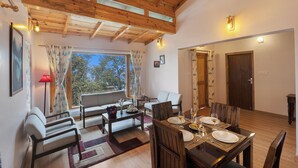 A dining area with a wooden table set for two, a sofa, and a window with curtains. - Alaya Stay Hill Home Petfriendly Cottage (Kosiyakutoli)