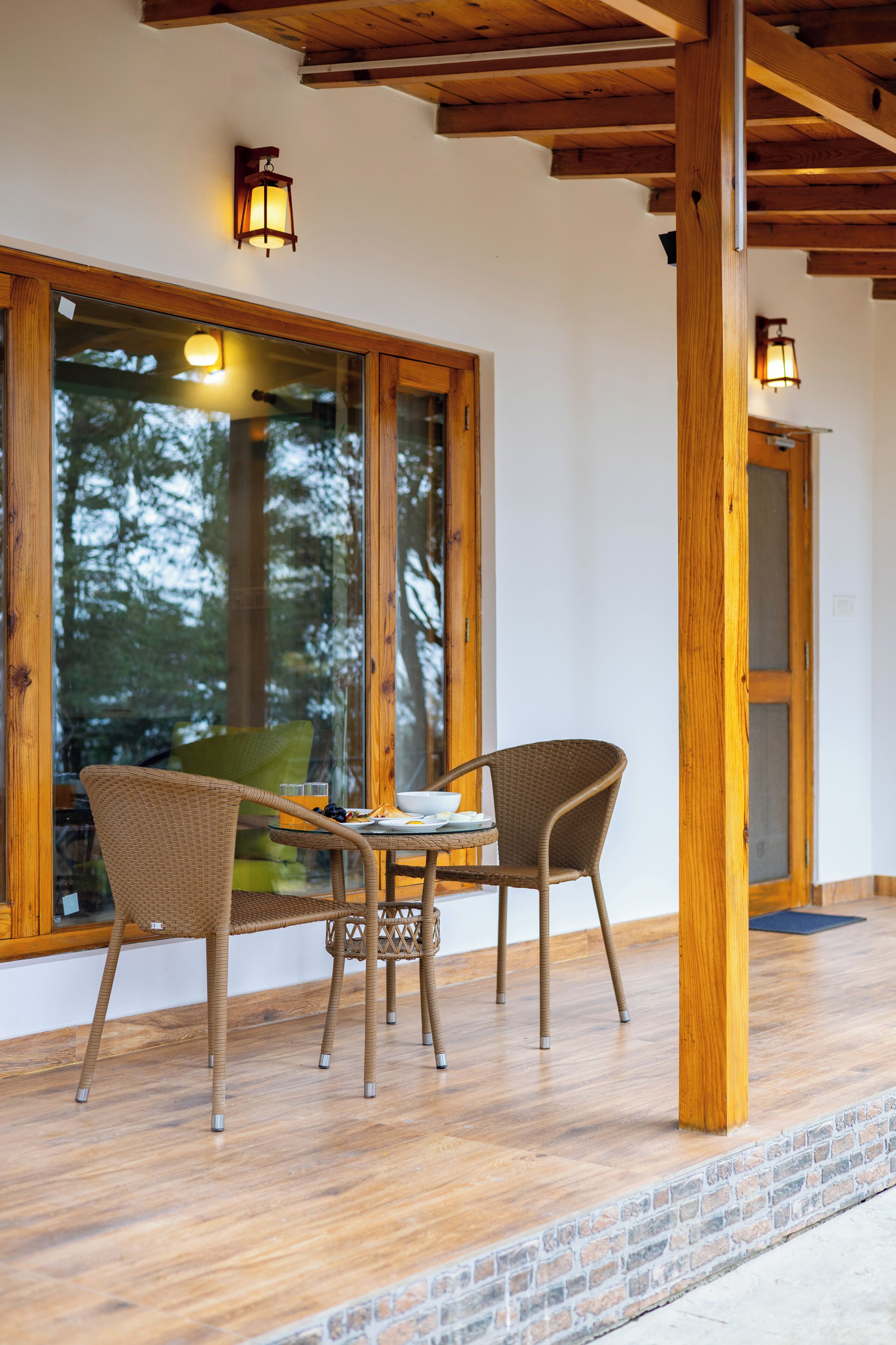 Una habitación con estructura de madera, puerta de cristal, sillas de mimbre, una mesa y dos luces fijadas a la pared.