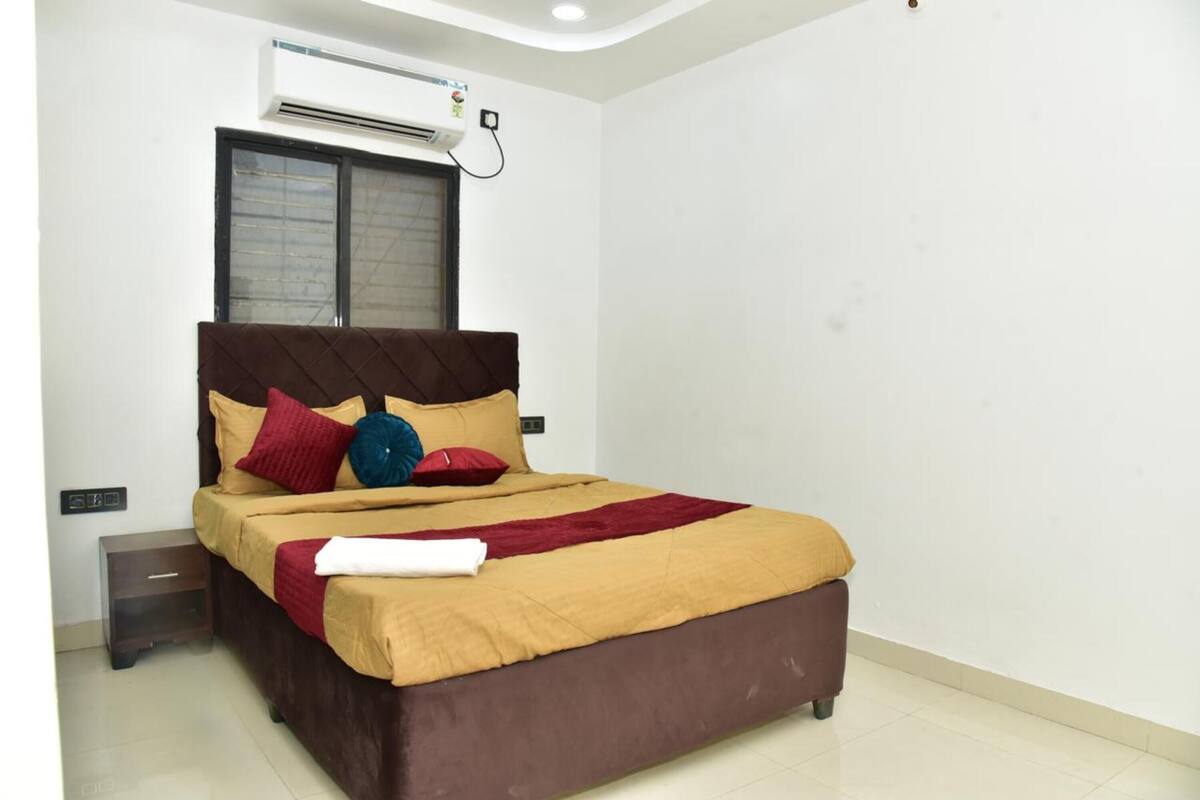 A bedroom with a bed, a nightstand, an air conditioning unit, and a window with blinds.