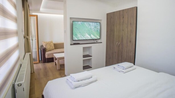 A modern hotel room with a bed, a TV mounted on the wall, a wooden wardrobe, and a seating area with a sofa and a table.