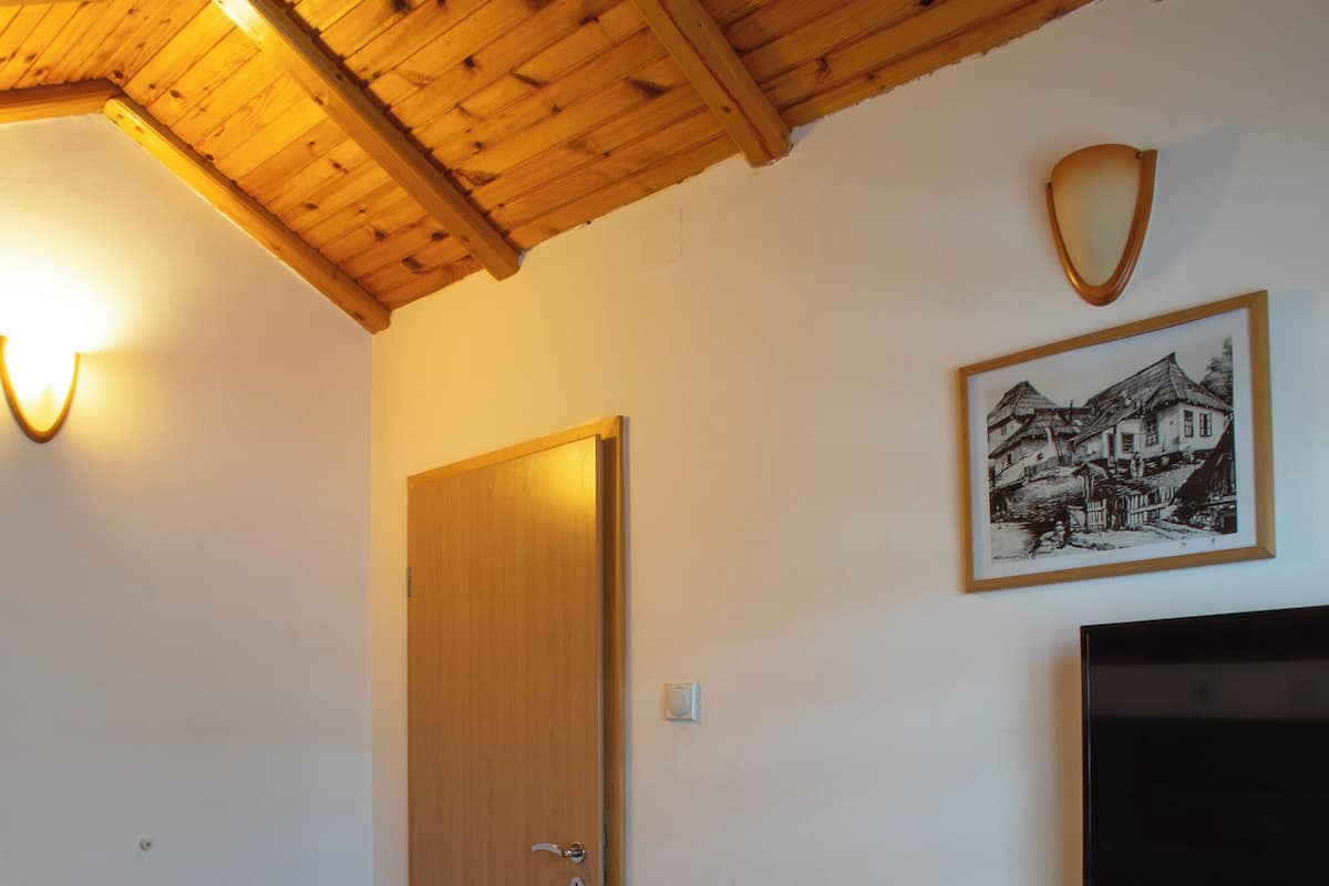 Une chambre d’hôtel avec un plafond en bois, un lit recouvert d’une couette blanche, une porte en bois, un tableau encadré au mur et un téléviseur à écran plat fixé au-dessus du lit.