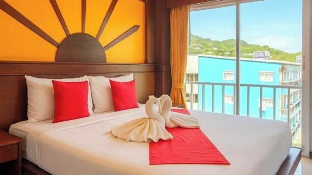 A hotel room with a large bed, a sun-shaped wall decoration, a view of the city, and a balcony.