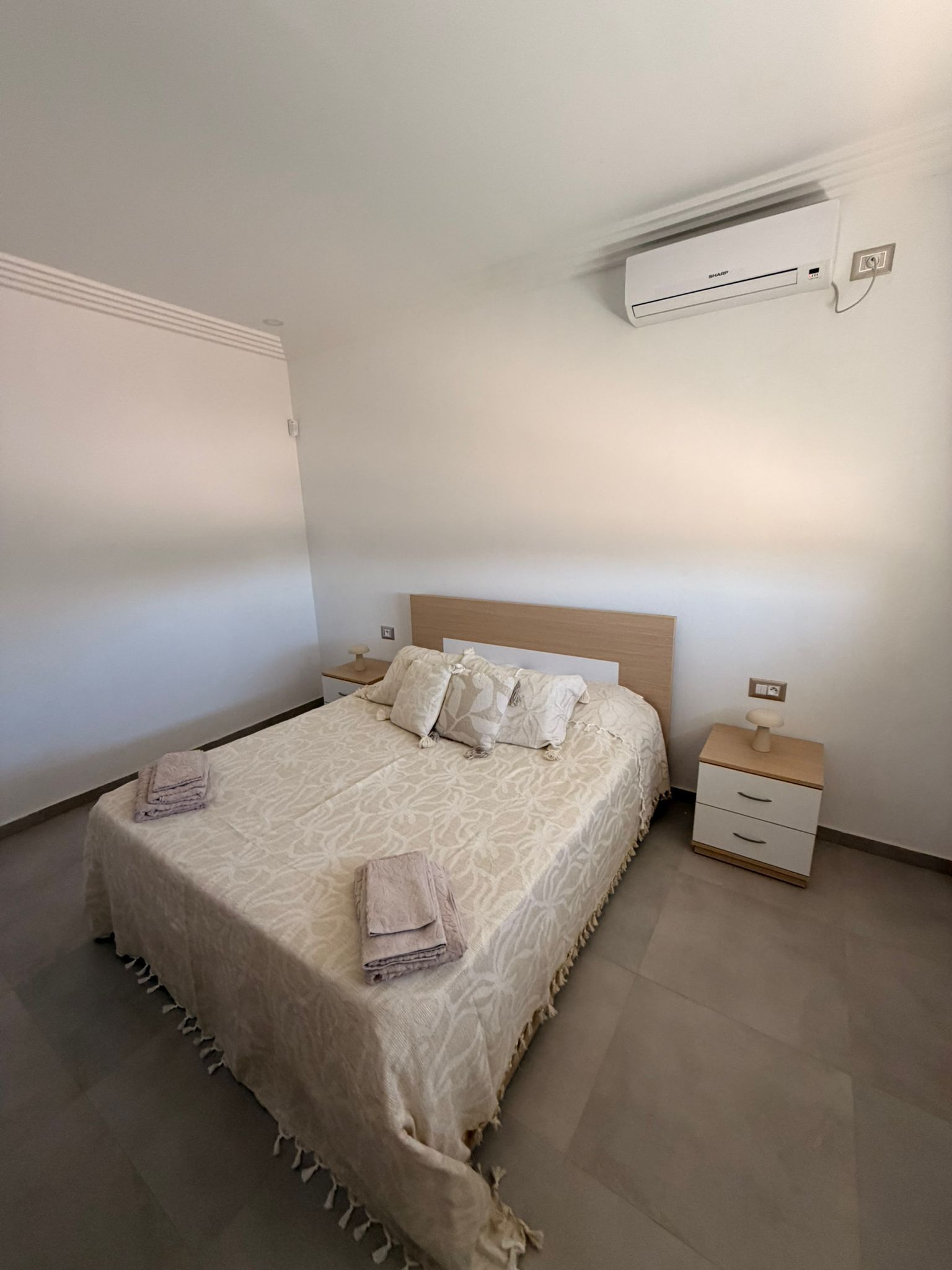 A neatly made bed with a patterned bedspread and pillows, a bedside table with a lamp, and an air conditioning unit mounted on the wall.