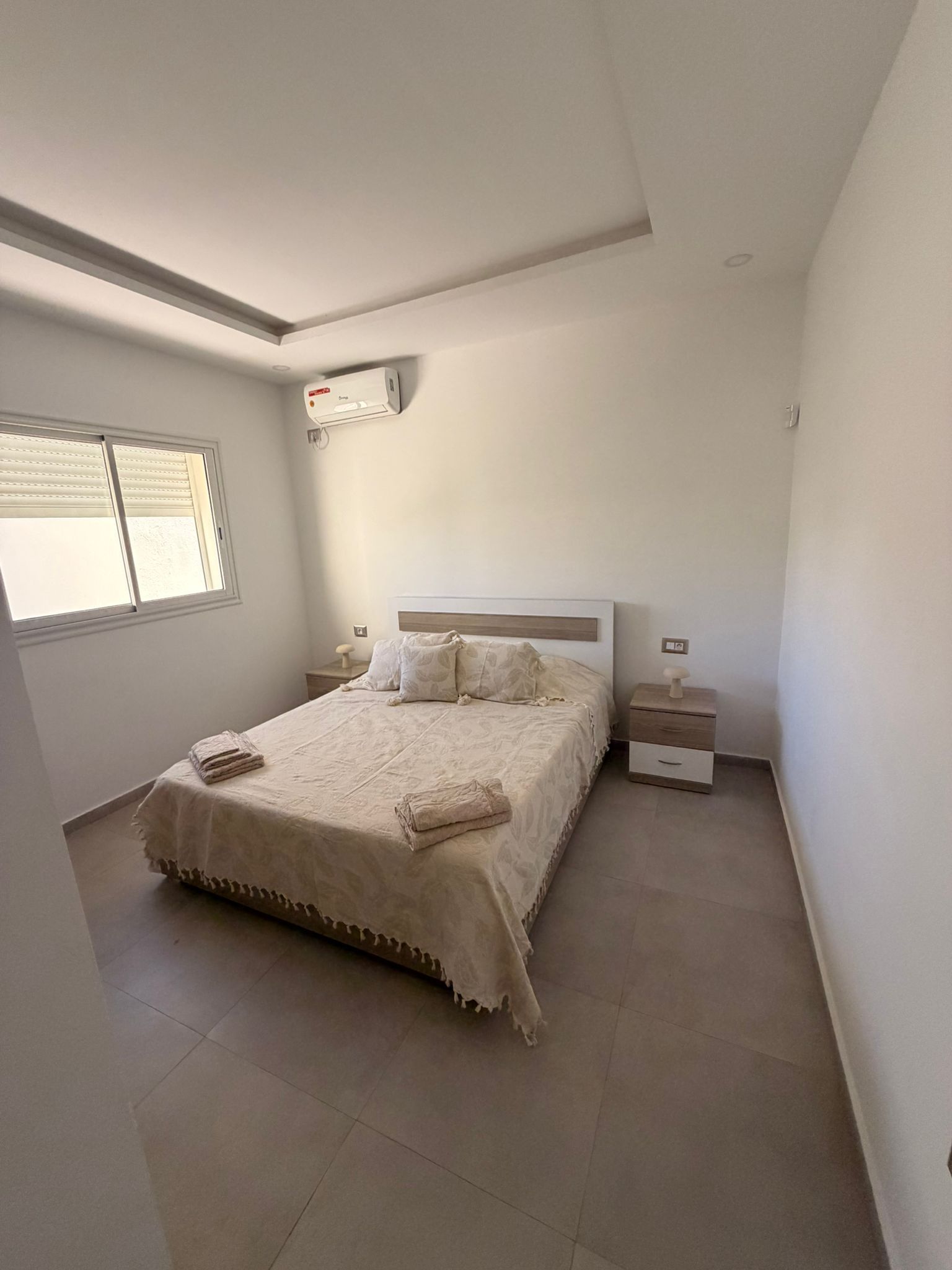 Une chambre à coucher comprenant un lit, des tables de chevet, un appareil de climatisation et une fenêtre.