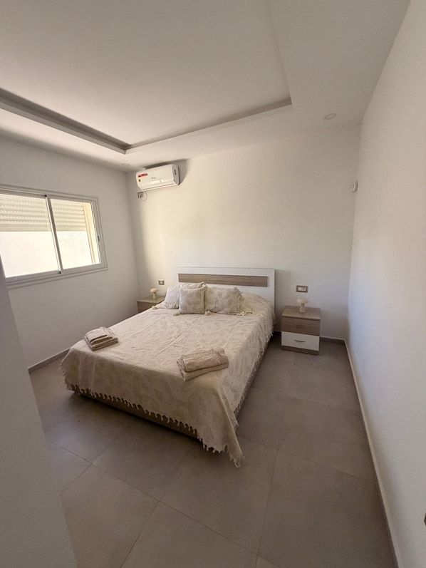 A bedroom with a bed, bedside tables, an air conditioning unit, and a window. - YASSMINE Villa (Djerba Midun)
