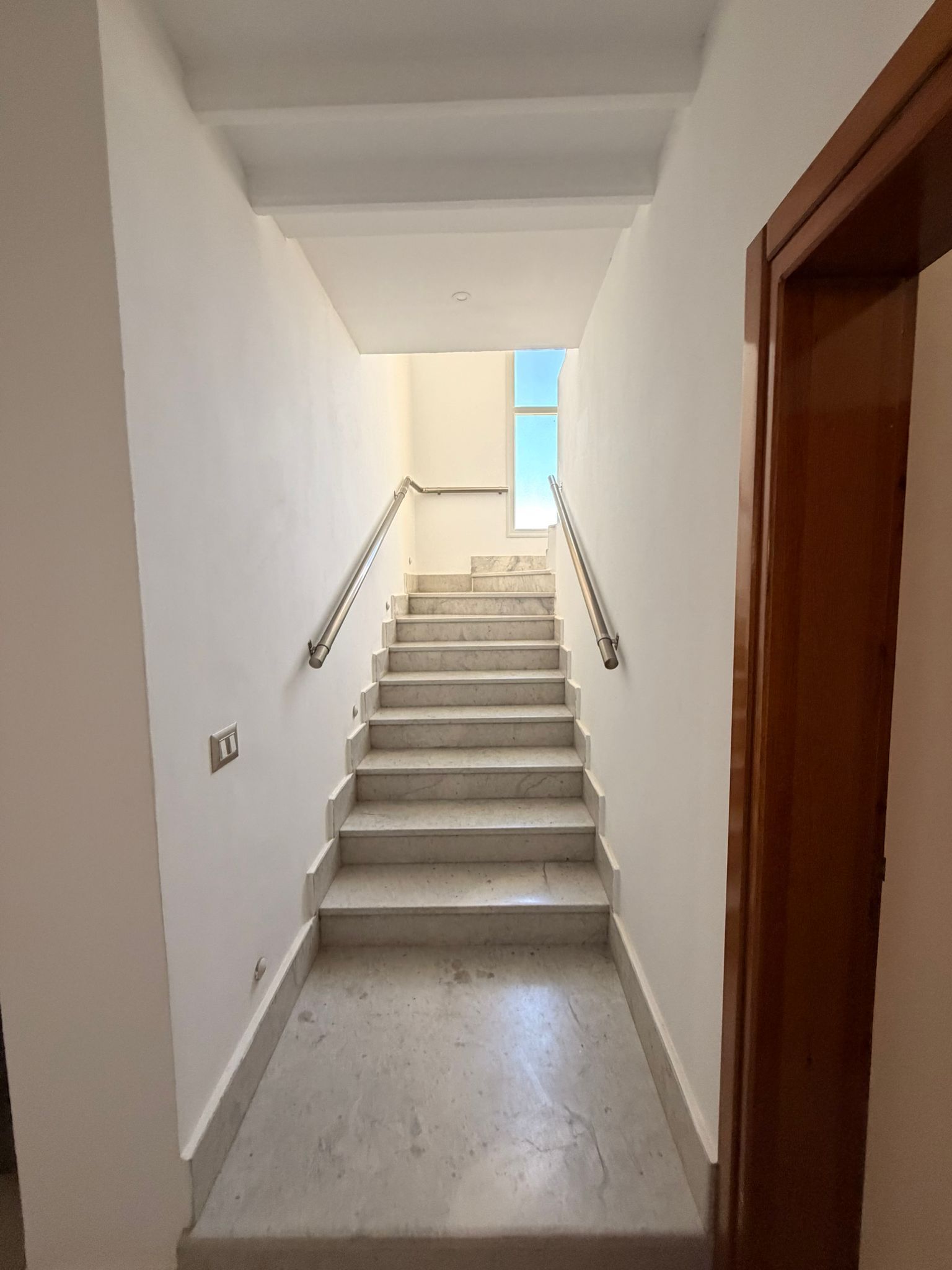 Uma escada com corrimão leva a um patamar, com uma porta à direita e uma janela no topo.