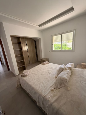 A bedroom with a large bed, built-in wardrobe, and a window with a view of greenery. - YASSMINE Villa (Djerba Midun)