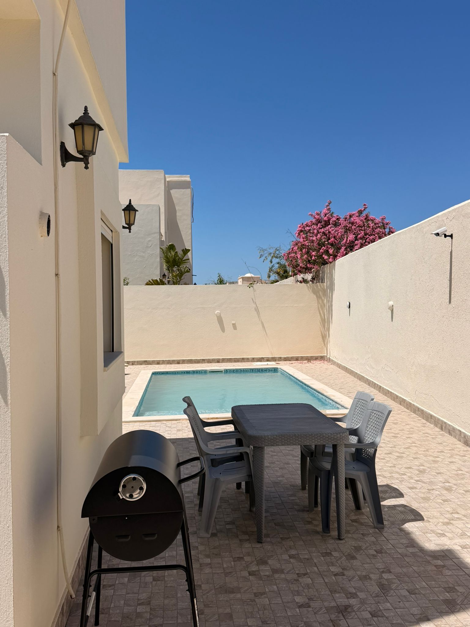 Une piscine avec une table et des chaises, un barbecue et une caméra de surveillance fixée au mur.
