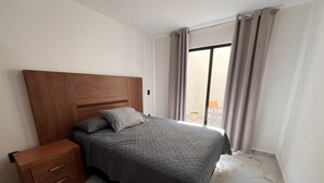 Ein Schlafzimmer mit einem Holz-Kopfteil, einem Bett mit grauer Steppdecke, einem Nachttisch mit Schubladen und Blick ins Freie durch eine Schiebetür.