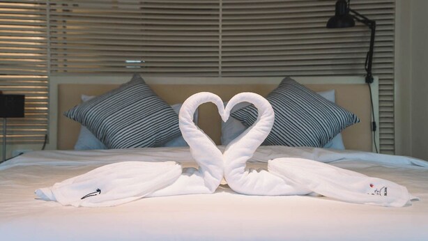 Habitación de hotel con cama, almohadas a rayas, una toalla dispuesta en forma de corazón y una lámpara.