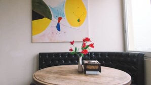 Una mesa redonda de madera con un jarrón de flores rojas y un libro, un sofá de cuero negro y un cuadro abstracto y colorido en la pared.