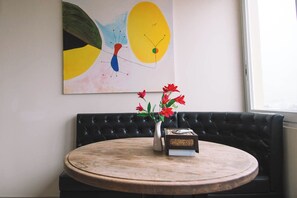 A round wooden table with a vase of red flowers and a book, a black leather sofa, and a colorful abstract painting on the wall. - Patong Penthouse 120m (Phuket)