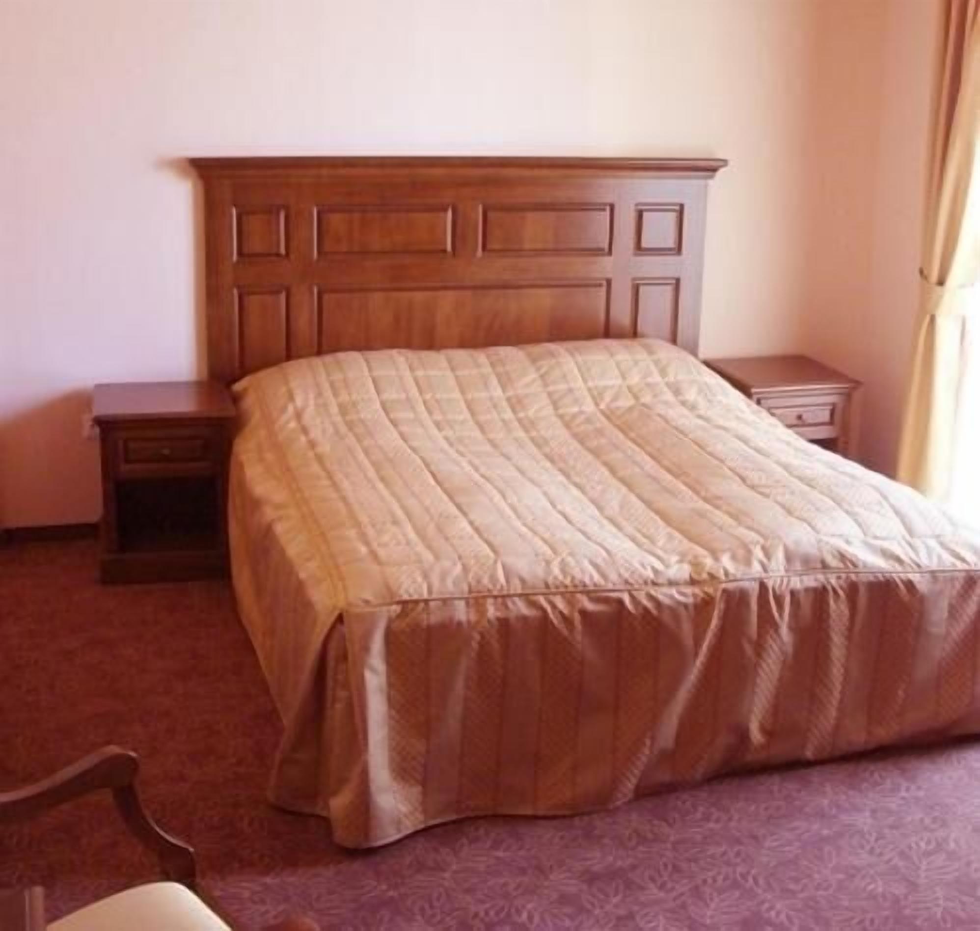 Una habitación de hotel con cabecera de madera, cama con colcha color beige, mesita de noche y cómoda.